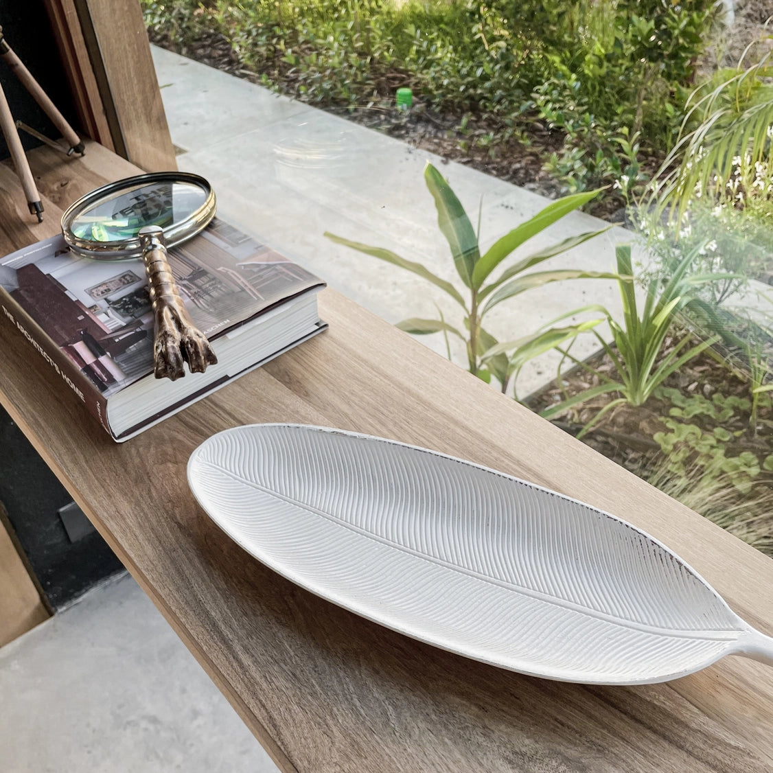 White ceramic leaf-shaped serving platter with embossed vein texture on natural wood shelf beside magnifying glass and books
