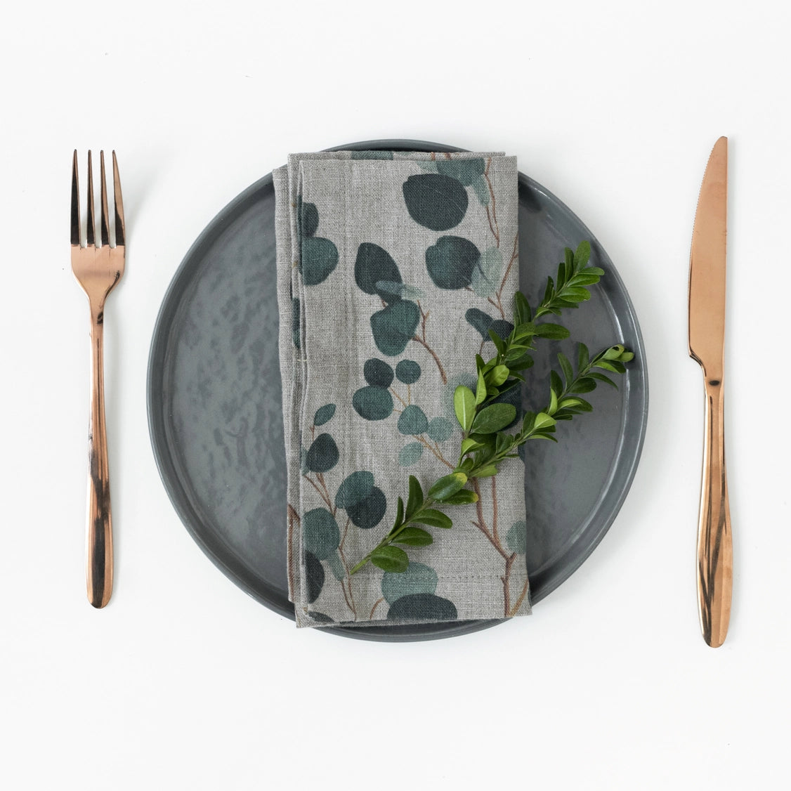 Natural linen napkin with dark green eucalyptus leaf print on charcoal plate, flanked by rose gold cutlery and greenery