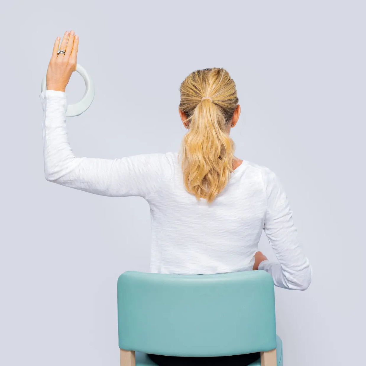 Blonde woman in white long-sleeve top seated on mint green round ottoman with wooden legs, back view on gray background