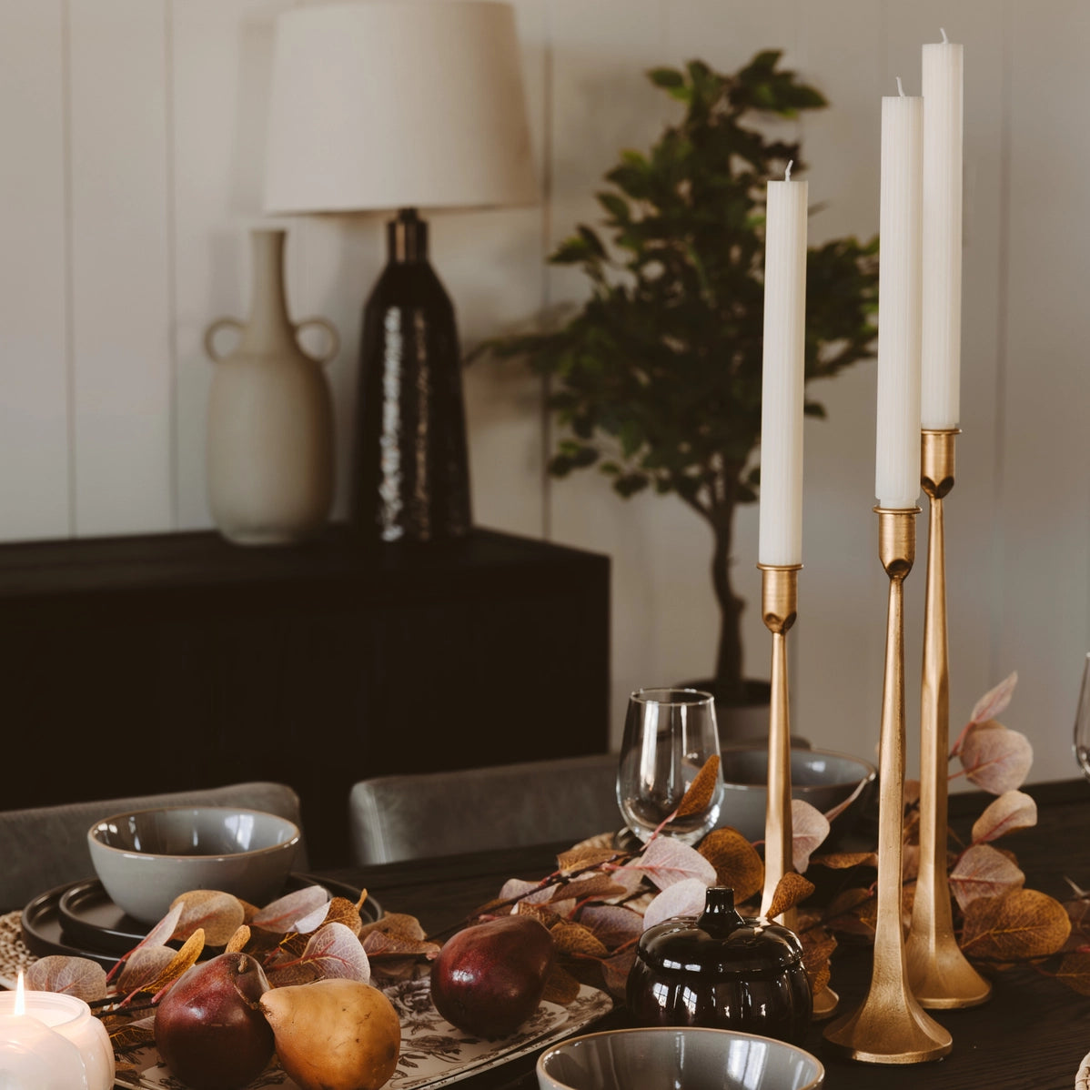 Brushed gold taper candlestick holders with white candles on autumn table with pears and dried eucalyptus leaves