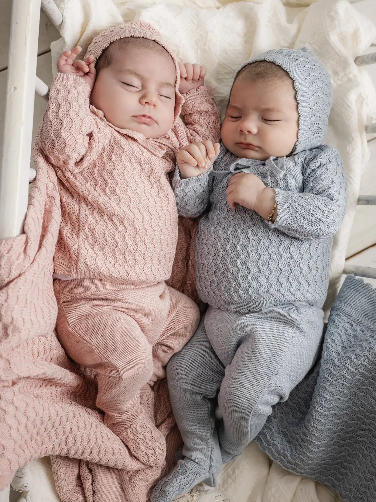 Knitted baby layette sets in dusty rose and sage blue with cable-knit texture, including hooded cardigans and matching pants
