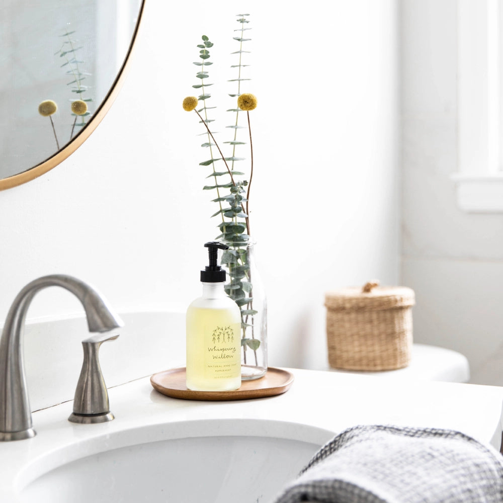 Clear glass soap dispenser with black pump and pale yellow liquid on wooden tray beside eucalyptus stems and dried flowers