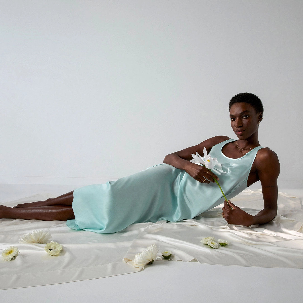 Model reclining in mint green sleeveless midi dress with fitted bodice holding white daisy on ivory fabric backdrop