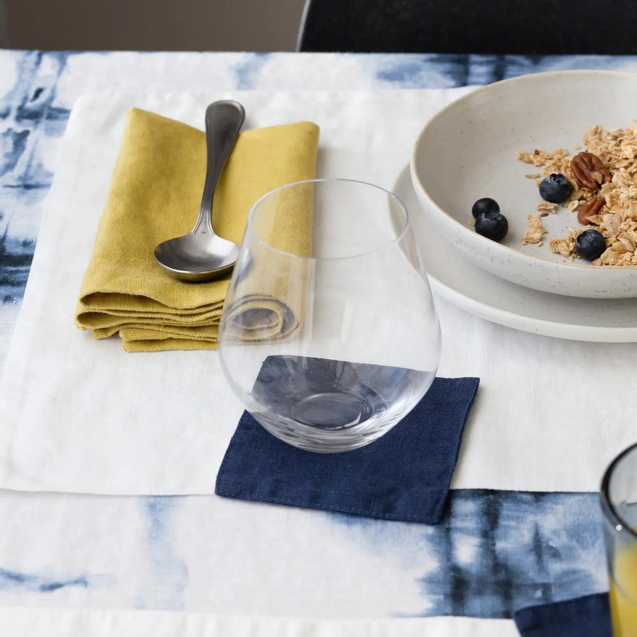 Stemless wine glass on navy linen coaster beside mustard yellow napkins and breakfast bowl with granola and blueberries
