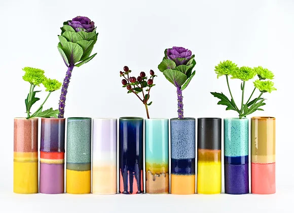 Row of ceramic cylinder vases with color-blocked glazes in metallic copper, pink, teal, navy, and gold holding purple cabbage and green flowers