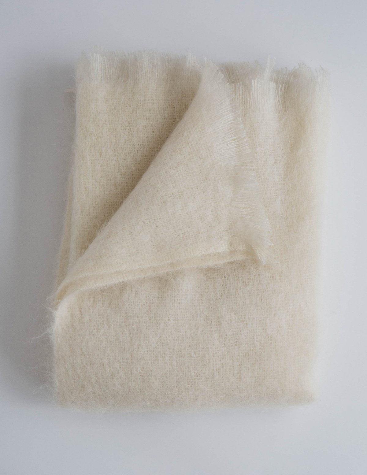 Cream mohair throw blanket with raw frayed edges, soft fuzzy texture, artfully folded showing lightweight weave