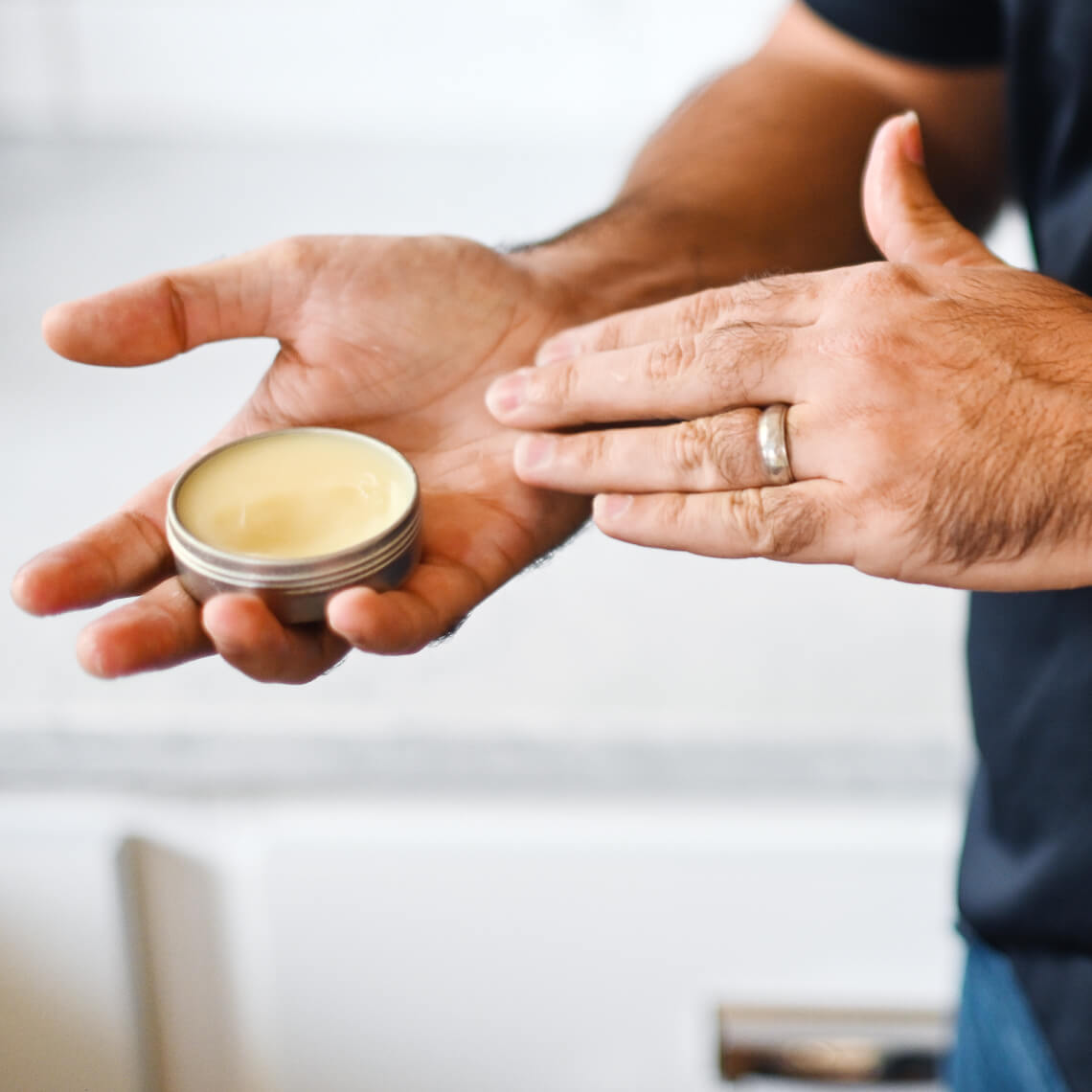 Hand applying pale yellow balm from small silver tin onto back of other hand wearing wedding band on bright background