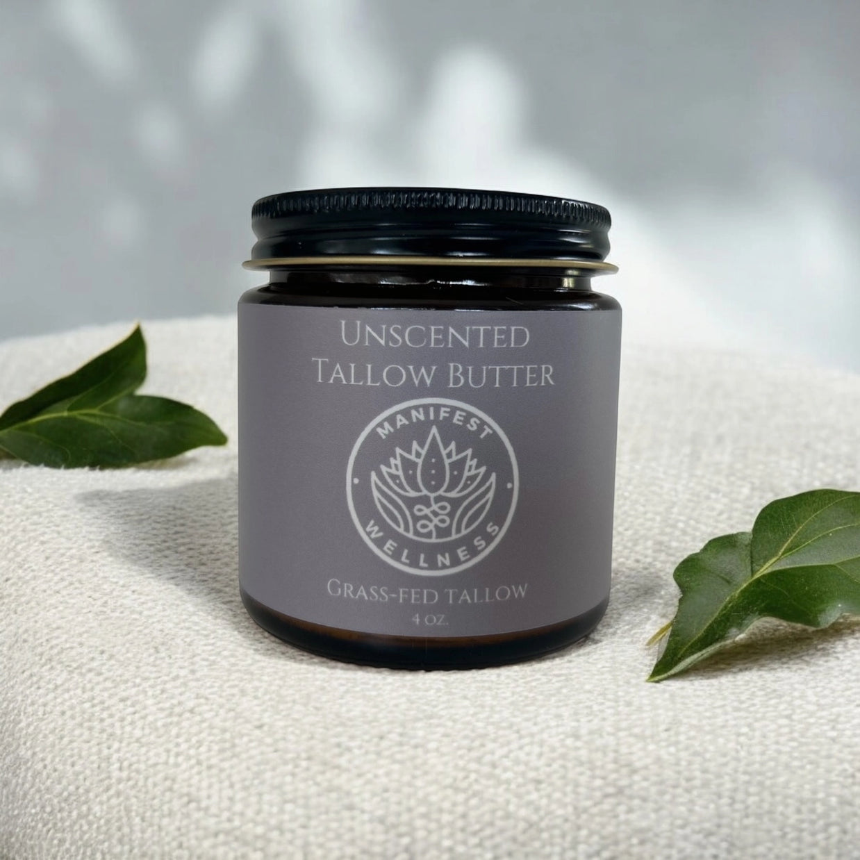 Amber glass jar with black lid containing unscented tallow butter with gray label and lotus logo on white textured fabric
