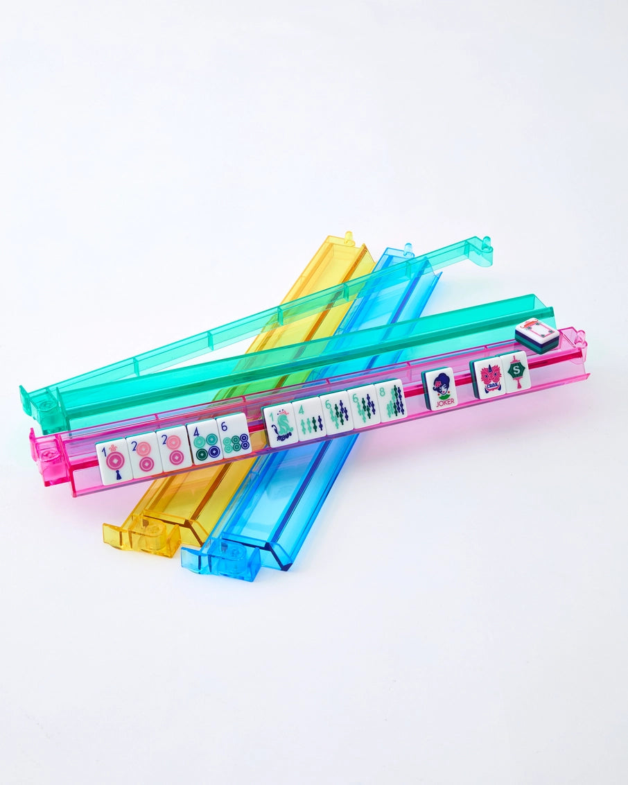 Transparent acrylic mahjong tile racks in pink, turquoise, yellow, and blue with white tiles displaying colorful dots and characters