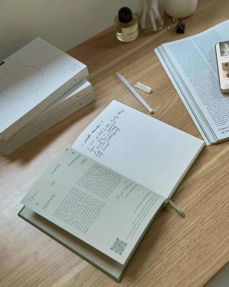 Light oak wood desk with open journals, white pen, stacked minimalist notebooks, perfume bottle and round candle