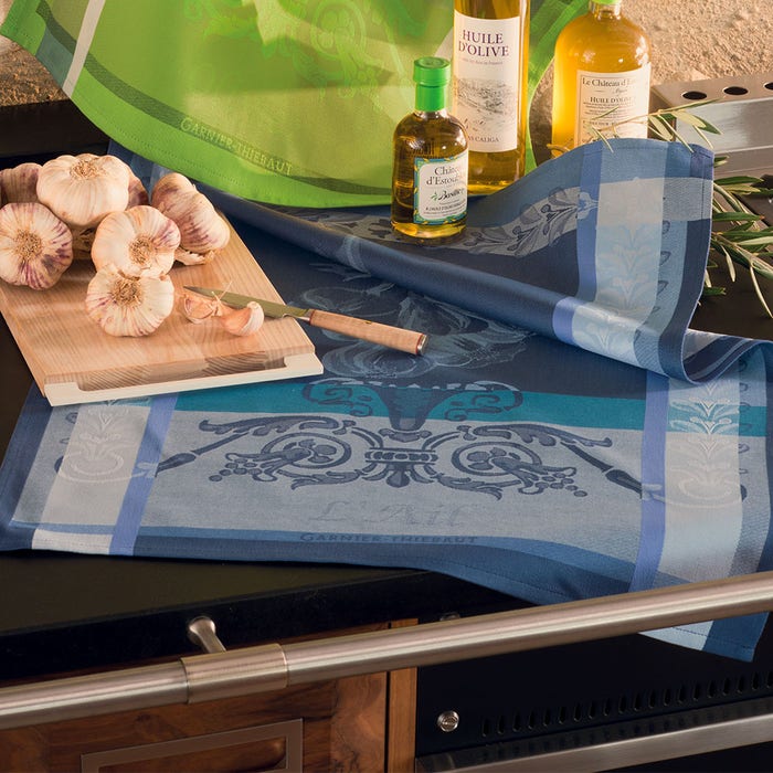 French jacquard kitchen towel in blue tones with ornate baroque scroll pattern alongside garlic, olive oil bottles, and cooking utensils