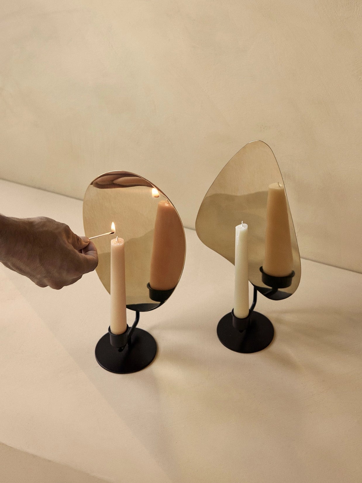 Modern candle holders with matte black base, white tapered candles, curved amber and clear glass shields on cream surface