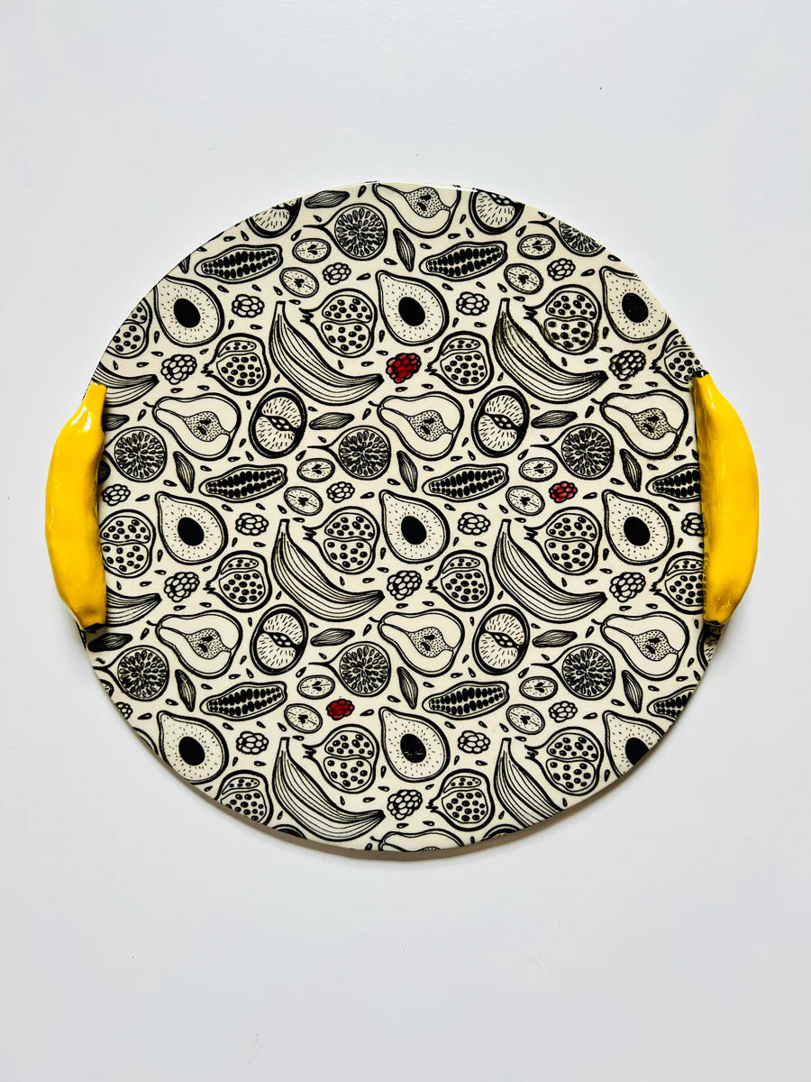 Round ceramic plate with black and white illustrated fruit pattern and bright yellow curved handles resembling banana peels
