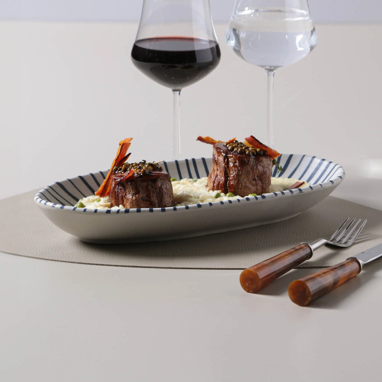 Braised short ribs with carrot garnish on white striped plate beside red and white wine glasses with wood-handled cutlery