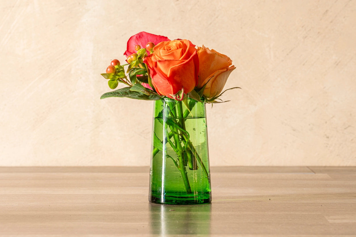 Coral and orange roses with pink ranunculus and green berries in emerald green glass vase on wood table