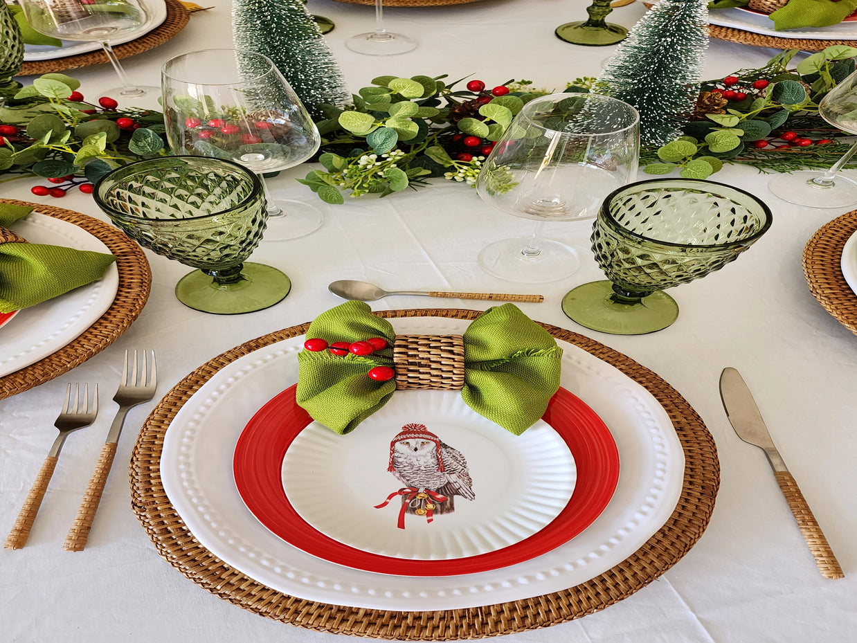 Holiday table setting with red-rimmed owl plates, green textured goblets, woven chargers, and frosted bottle brush trees