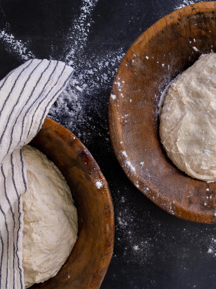 Round wooden dough bowls with risen bread dough dusted with flour on dark surface with striped kitchen towel