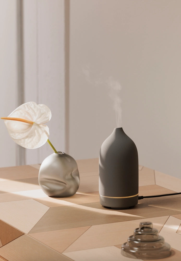 Matte gray ceramic essential oil diffuser with domed cap releasing mist beside round bud vase with white anthurium bloom