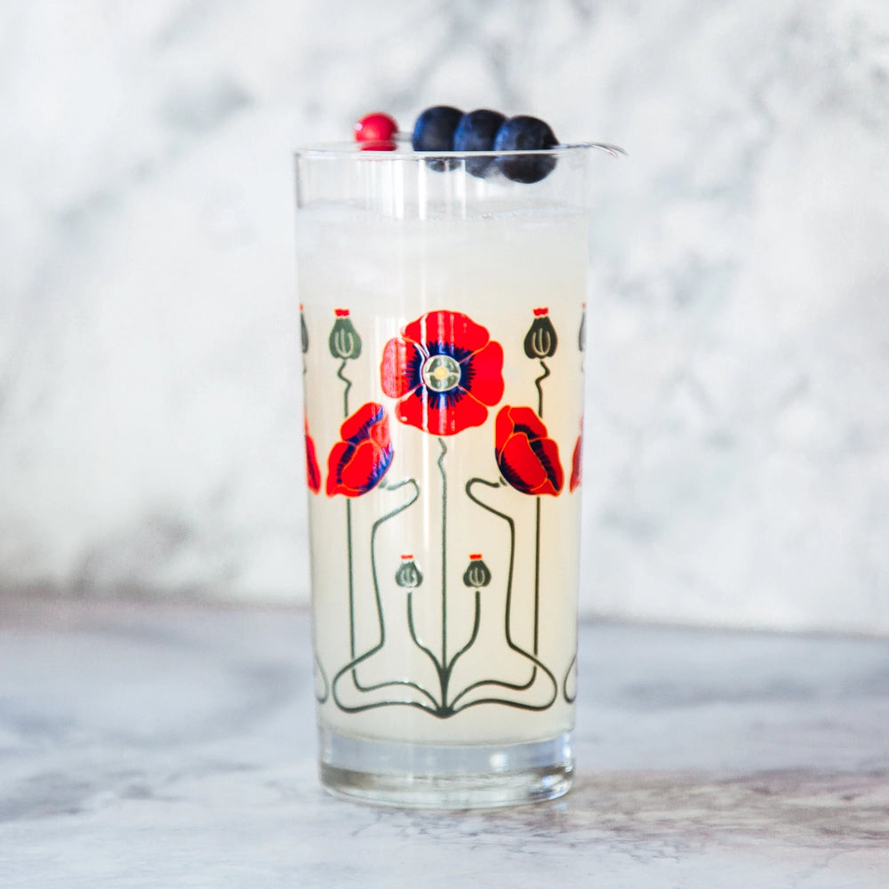 Clear glass tumbler with red poppy flower and green stem print, filled with white cocktail, garnished with blueberries