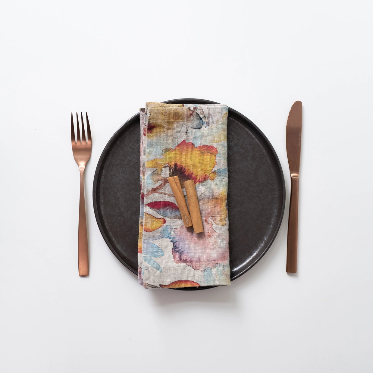 Linen napkin with watercolor floral pattern in orange, pink, yellow, and blue tones, topped with cinnamon sticks on black plate