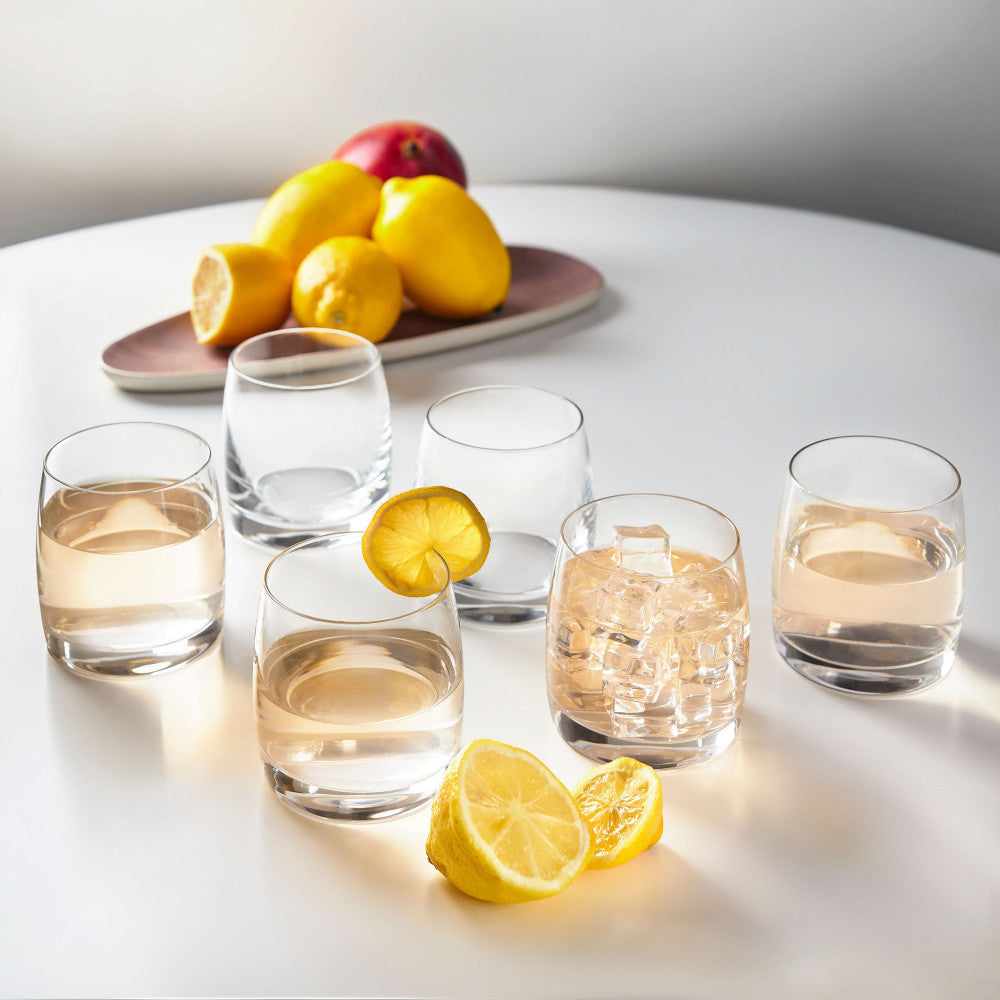 Set of six rounded clear glass tumblers with water and ice, fresh lemons and mango on dusty pink ceramic plate behind