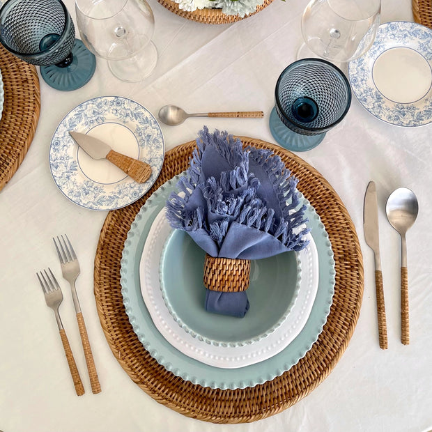 Coastal table setting with woven rattan chargers, blue floral china, sage green plates, fringed napkin, and textured teal goblets