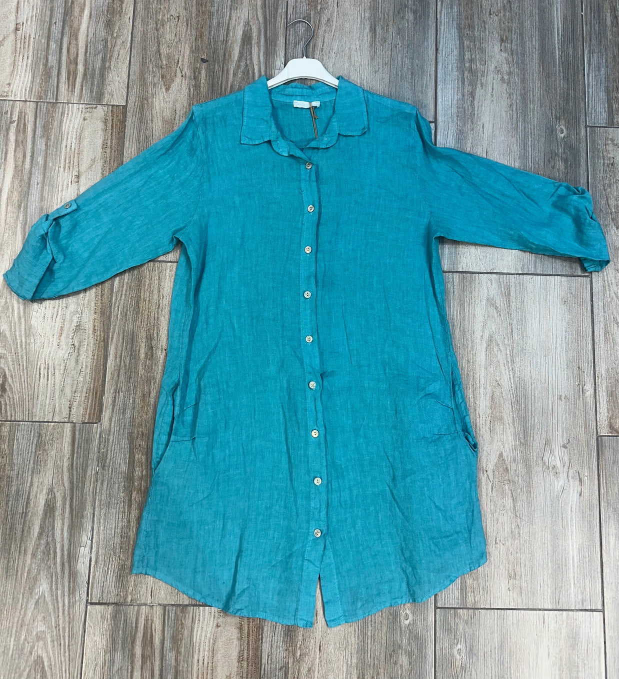Turquoise linen button-down shirt with rolled three-quarter sleeves and collar hanging on white hanger against weathered wood floor
