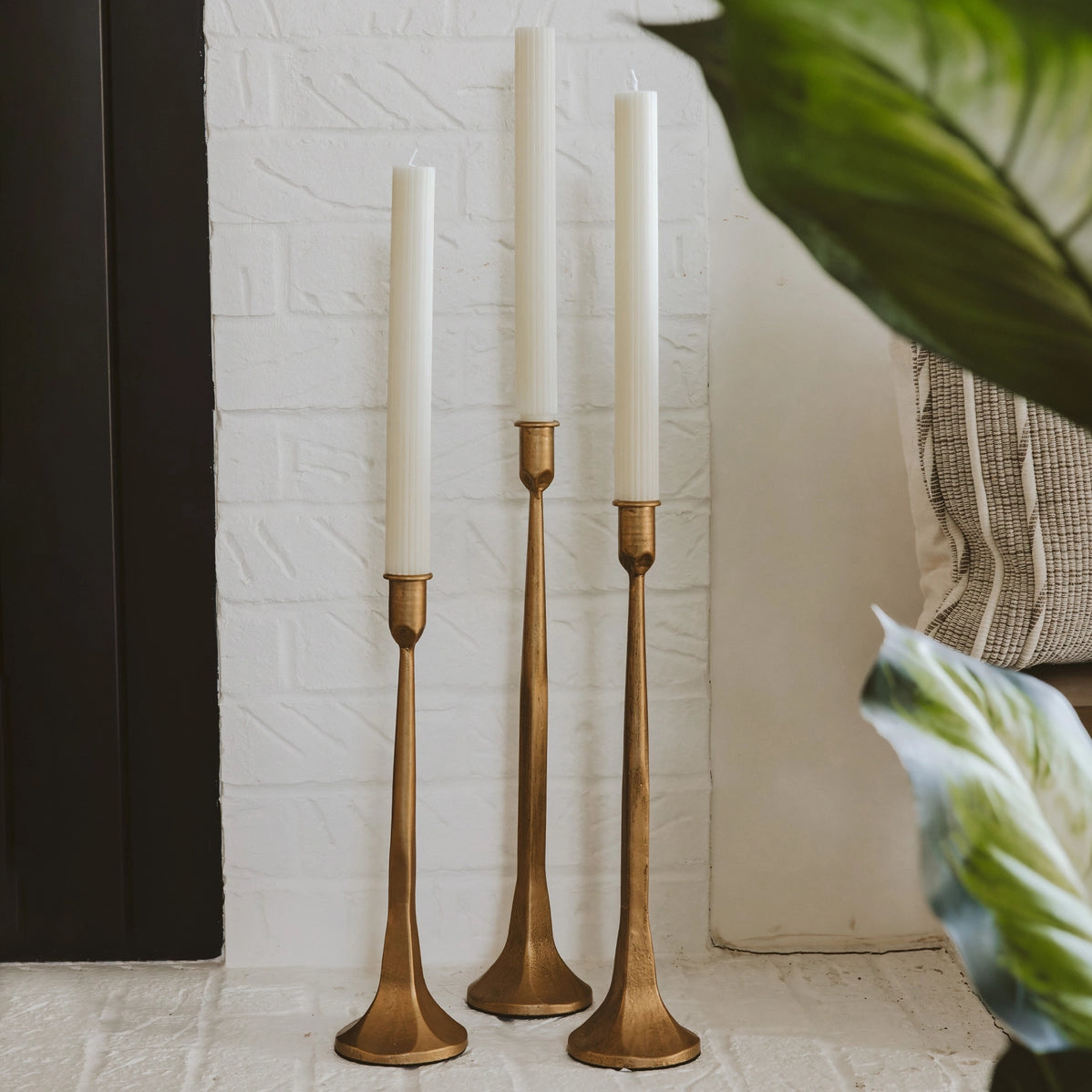 Three graduated brass taper candlesticks with long slender stems and flared bases holding white candles on stone surface