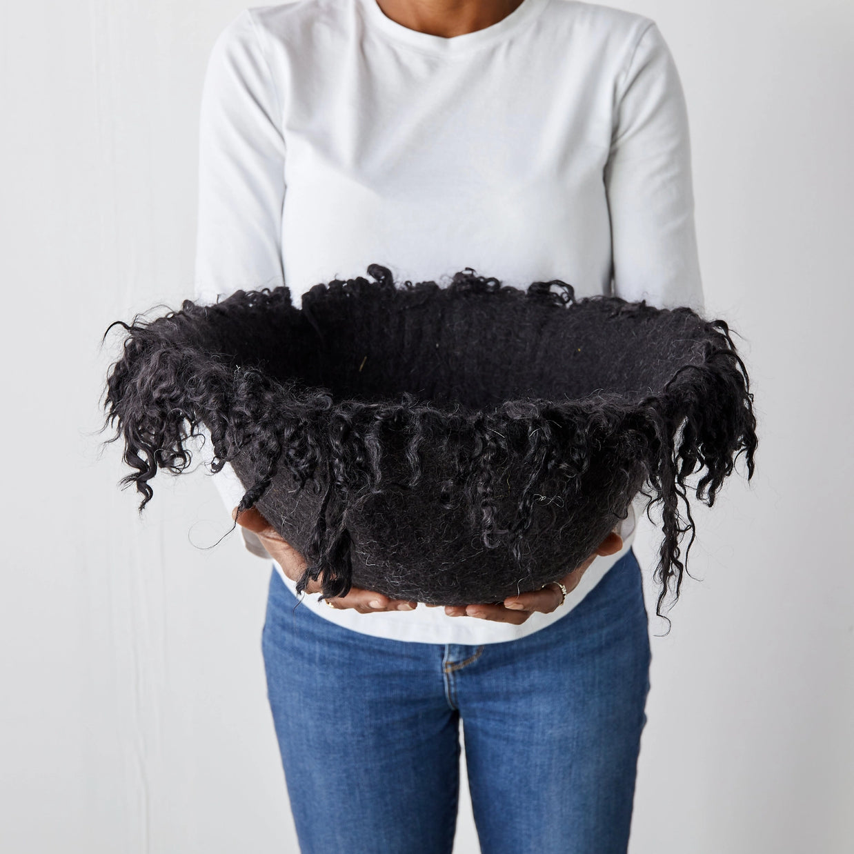 Black felted wool basket with long curly fringe trim around rim, held by person in white shirt against neutral background