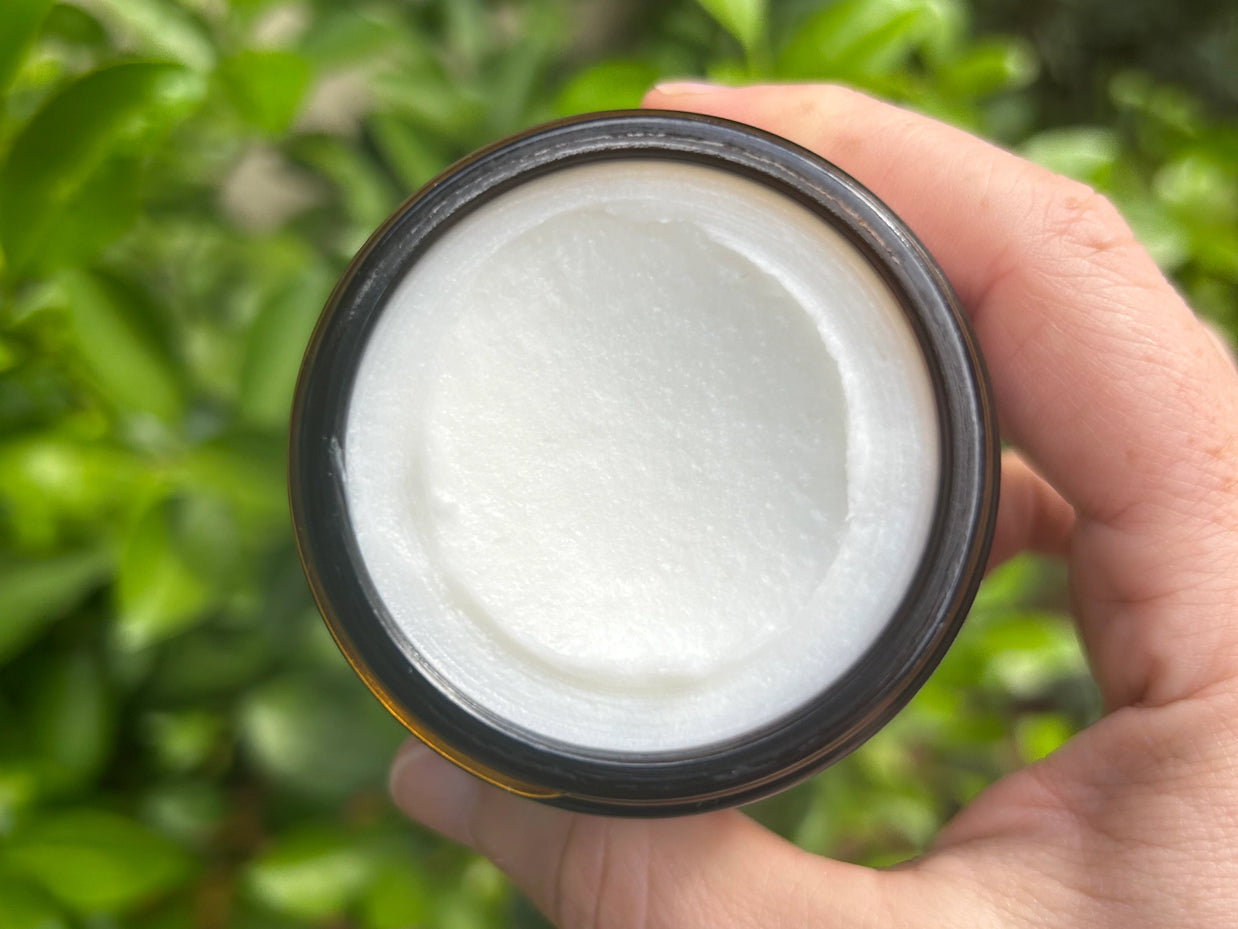 Open jar of white creamy moisturizer with smooth glossy texture in dark brown container held against blurred greenery