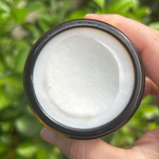 Open jar of white creamy moisturizer with smooth glossy texture in dark brown container held against blurred greenery