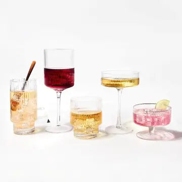 Assorted cocktail glassware collection with textured crystal, wine glass with burgundy liquid, and coupe glasses filled with amber and pink drinks