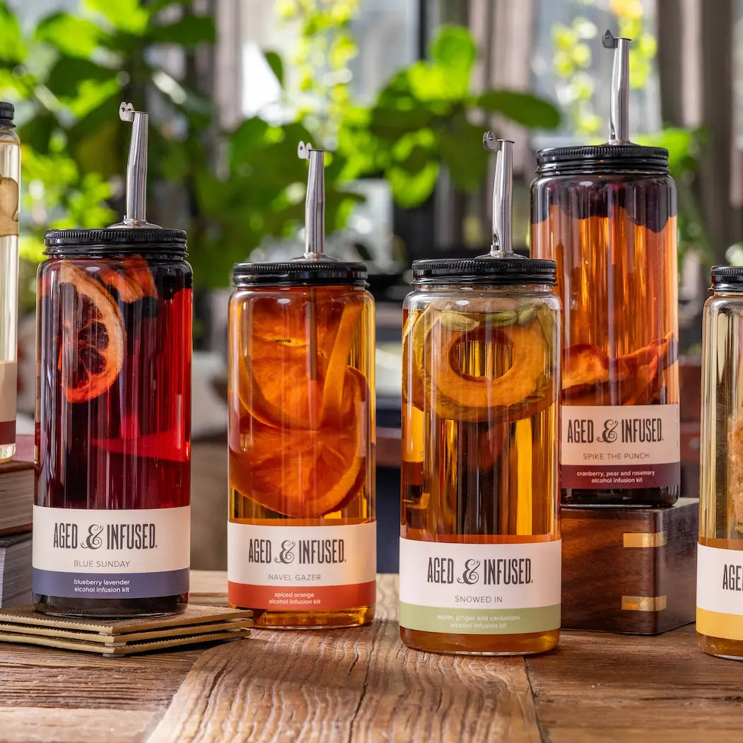 Glass mason jars with black lids and metal straws containing colorful fruit-infused alcohol mixtures in red, orange, and amber hues