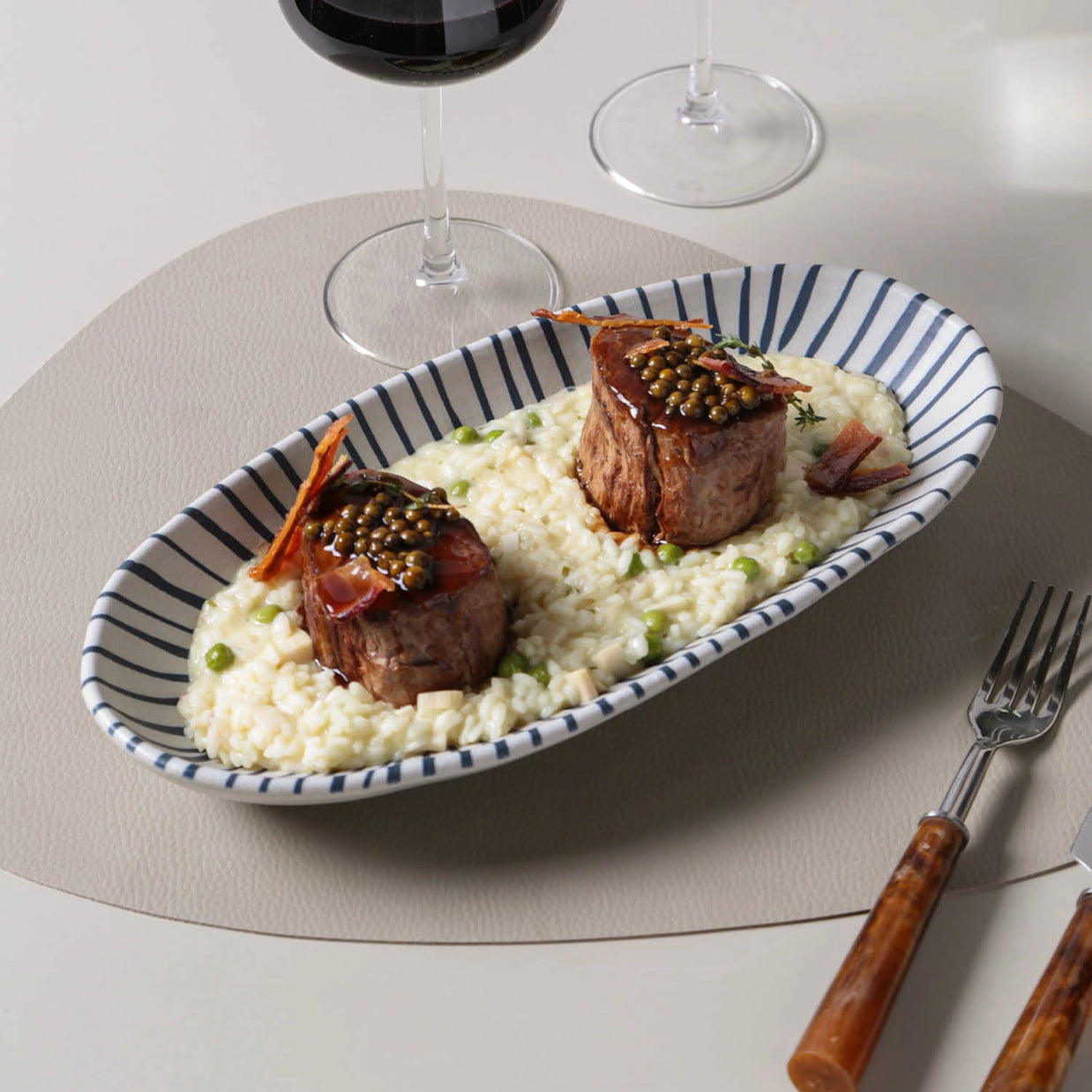 Oval ceramic serving platter with navy blue stripe pattern holding beef medallions over creamy risotto with peas and caviar garnish