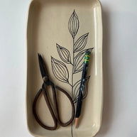 Cream ceramic rectangular catchall dish with black line drawing of botanical leaves holding small scissors and pen