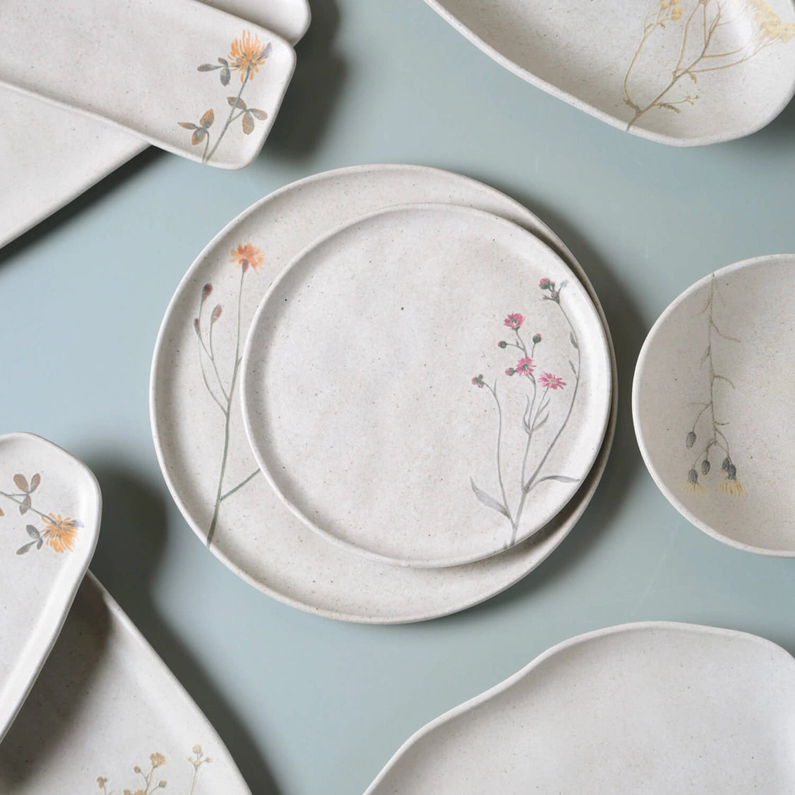 Handmade ceramic dinnerware set in speckled cream glaze with delicate hand-painted wildflower botanical designs on pale blue background