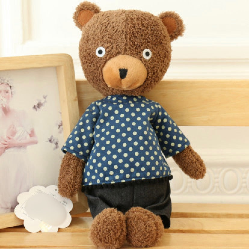 Brown plush teddy bear with tan muzzle wearing blue polka dot shirt and dark gray pants on wooden shelf