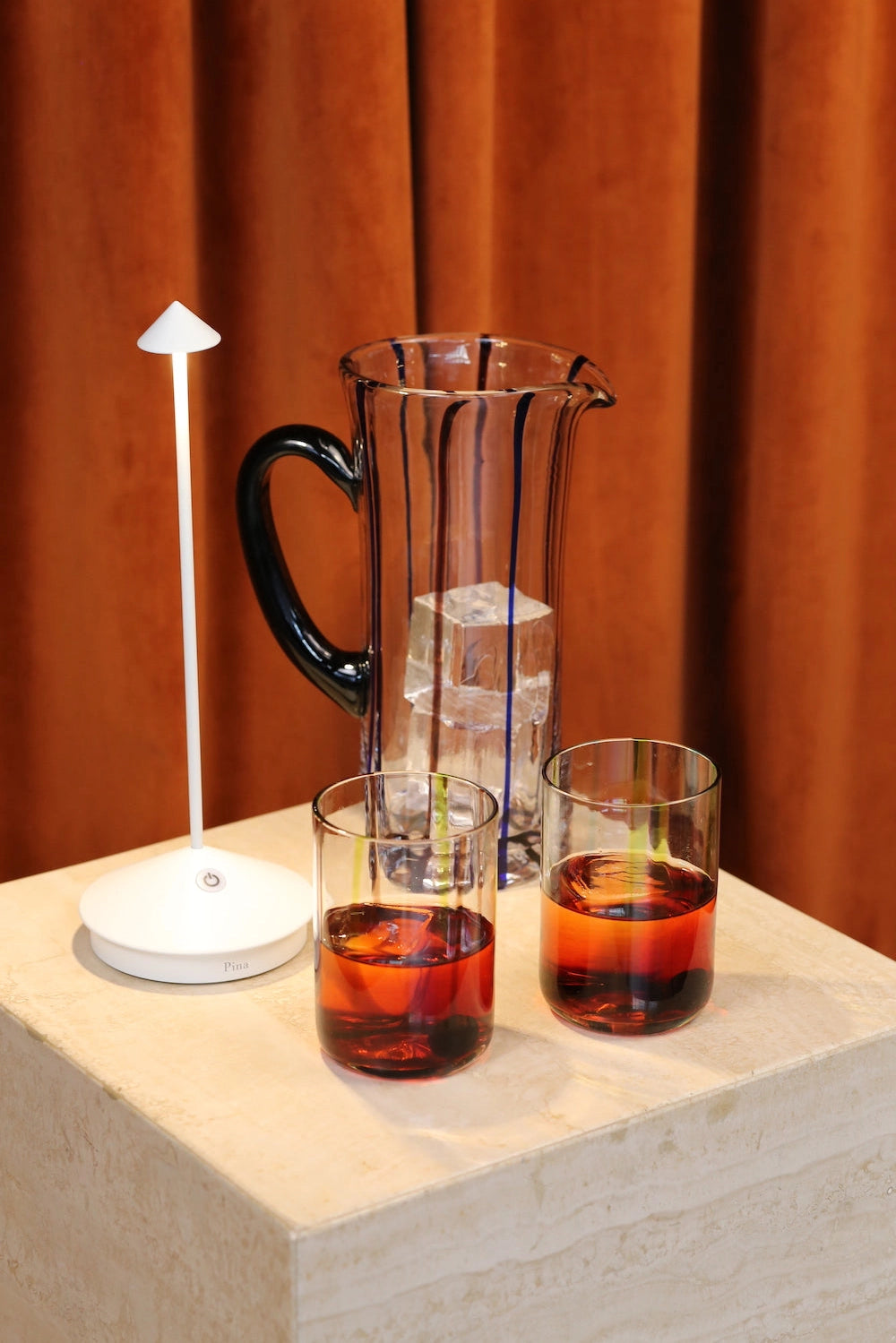 Clear glass pitcher with black handle and blue stripes, two amber cocktails with ice, white lamp on stone pedestal
