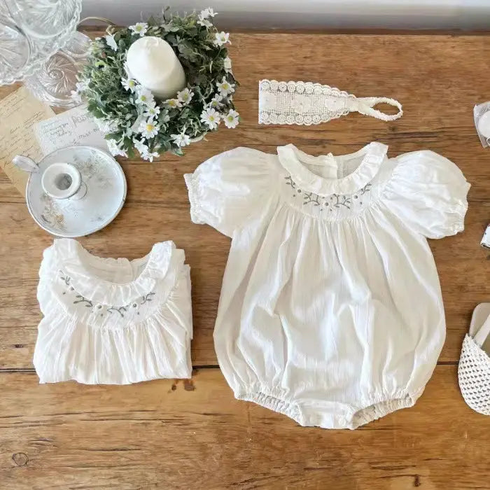 White cotton baby romper and dress with smocked embroidered necklines displayed on wood table with vintage accessories