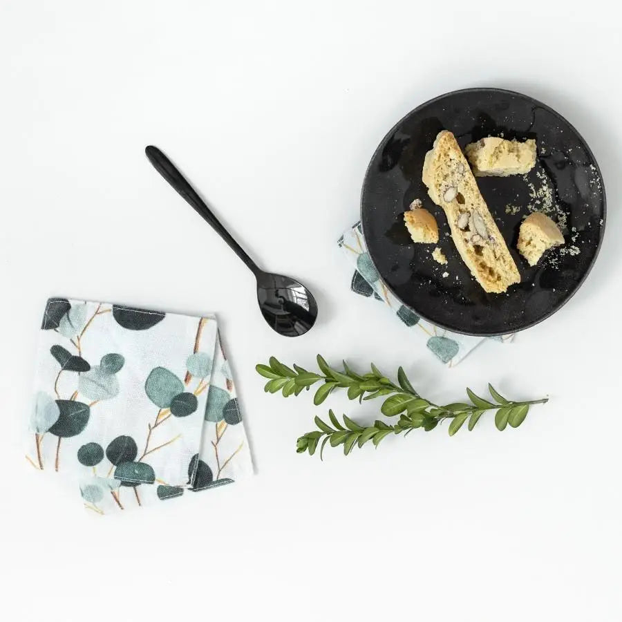 Set of white linen coasters with watercolor eucalyptus leaf pattern in sage and teal tones, displayed beneath black plate
