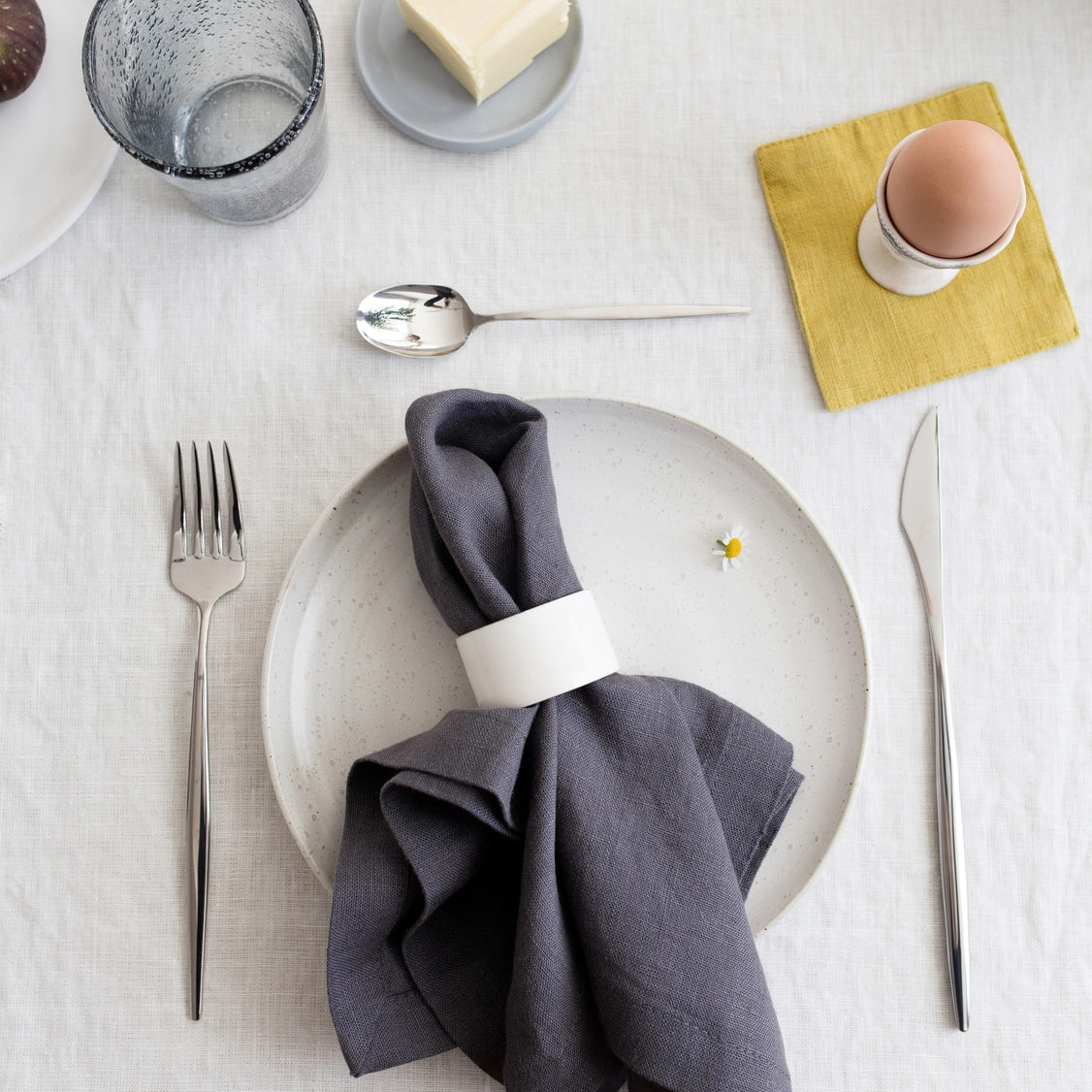 Minimalist breakfast table setting with speckled ceramic plate, charcoal linen napkin with white ring, and yellow cloth napkin