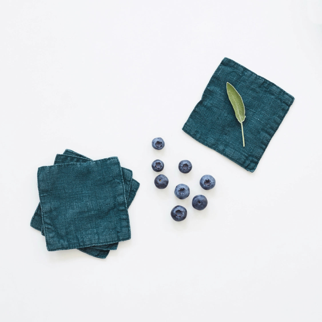 Set of four deep teal linen cocktail napkins with visible texture, styled with fresh blueberries and sage leaf