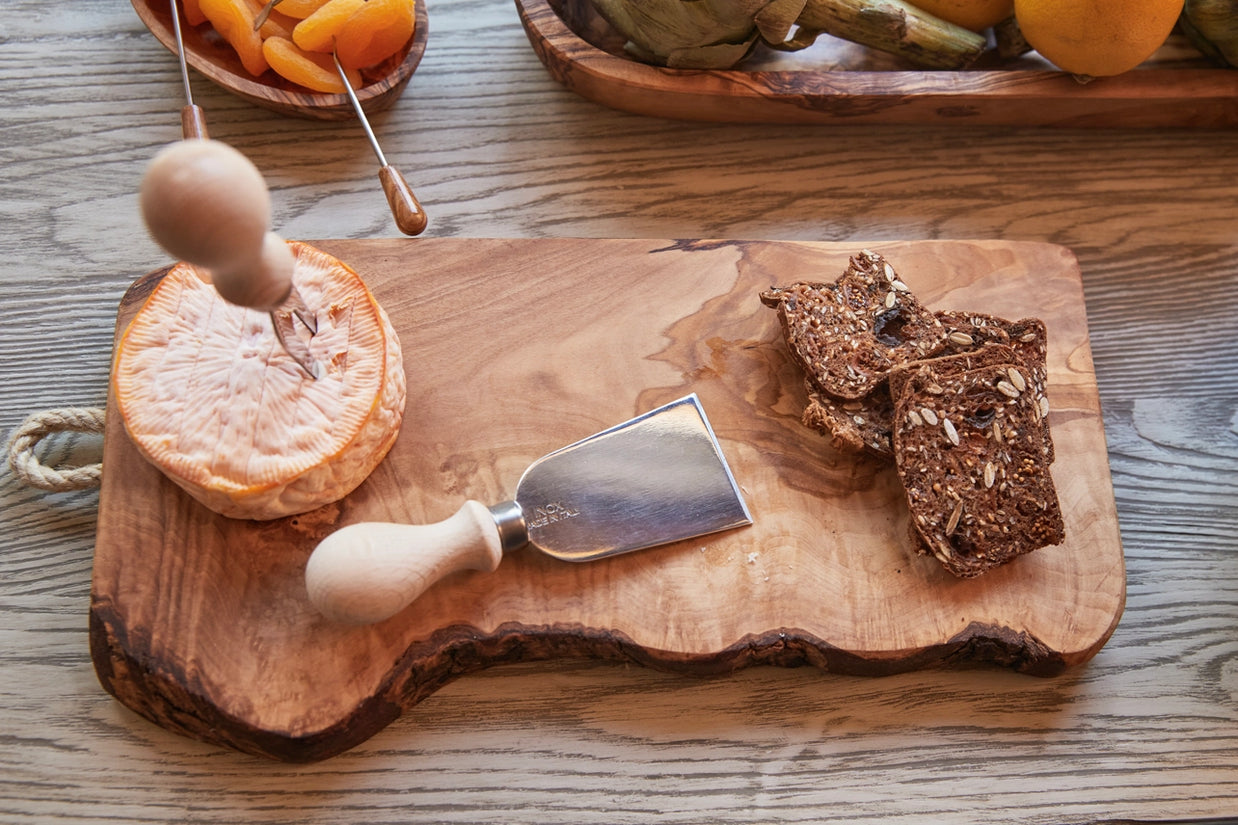 Live edge wooden serving board with cheese knife, seeded bread slices, and woven cheese basket on weathered wood table