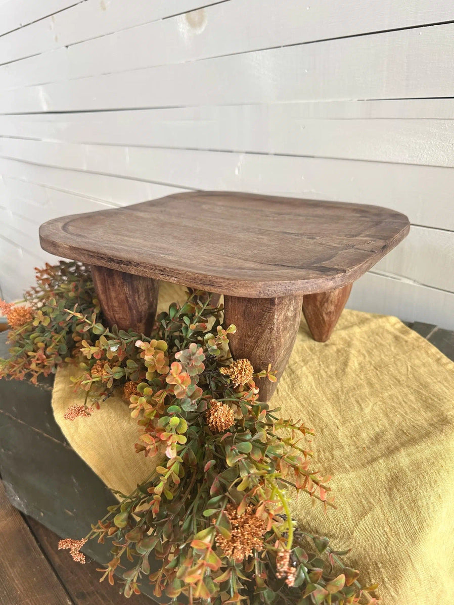 Rustic wooden pedestal stand with rounded square top and arched legs on yellow linen with autumn eucalyptus wreath