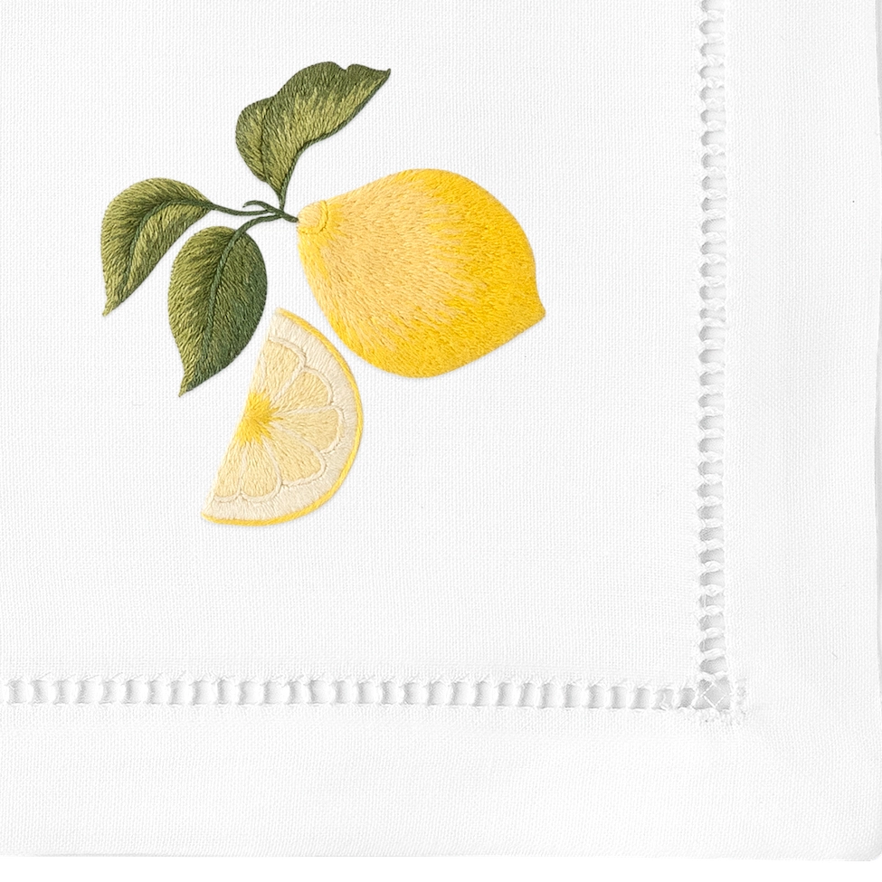 Embroidered white linen napkin with yellow lemon, cross-section slice, and olive green leaves in satin stitch detail