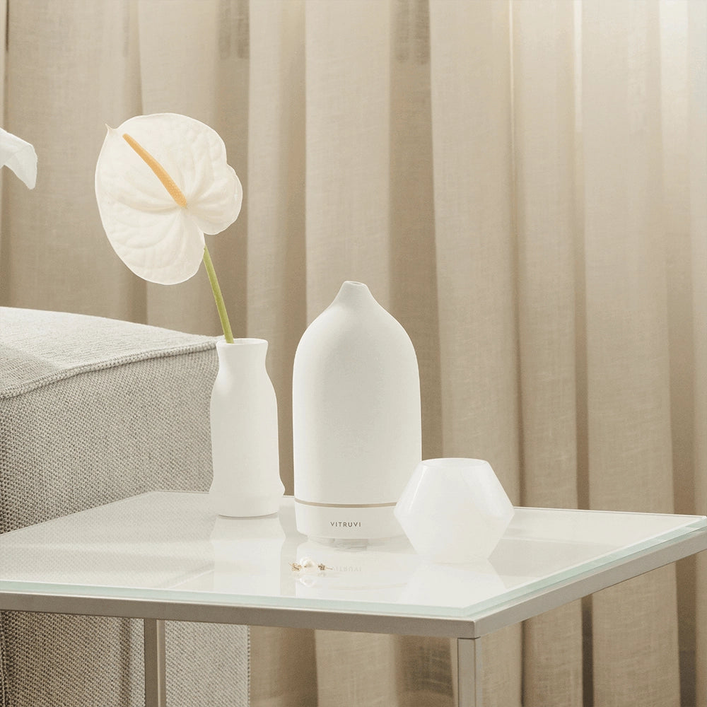 White matte ceramic diffuser with dome-shaped top and geometric hexagonal base beside white anthurium in bud vase on glass table