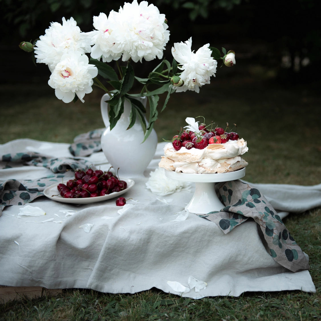 White ceramic pitcher with fluffy white peonies beside layered pavlova topped with fresh berries on marble cake stand