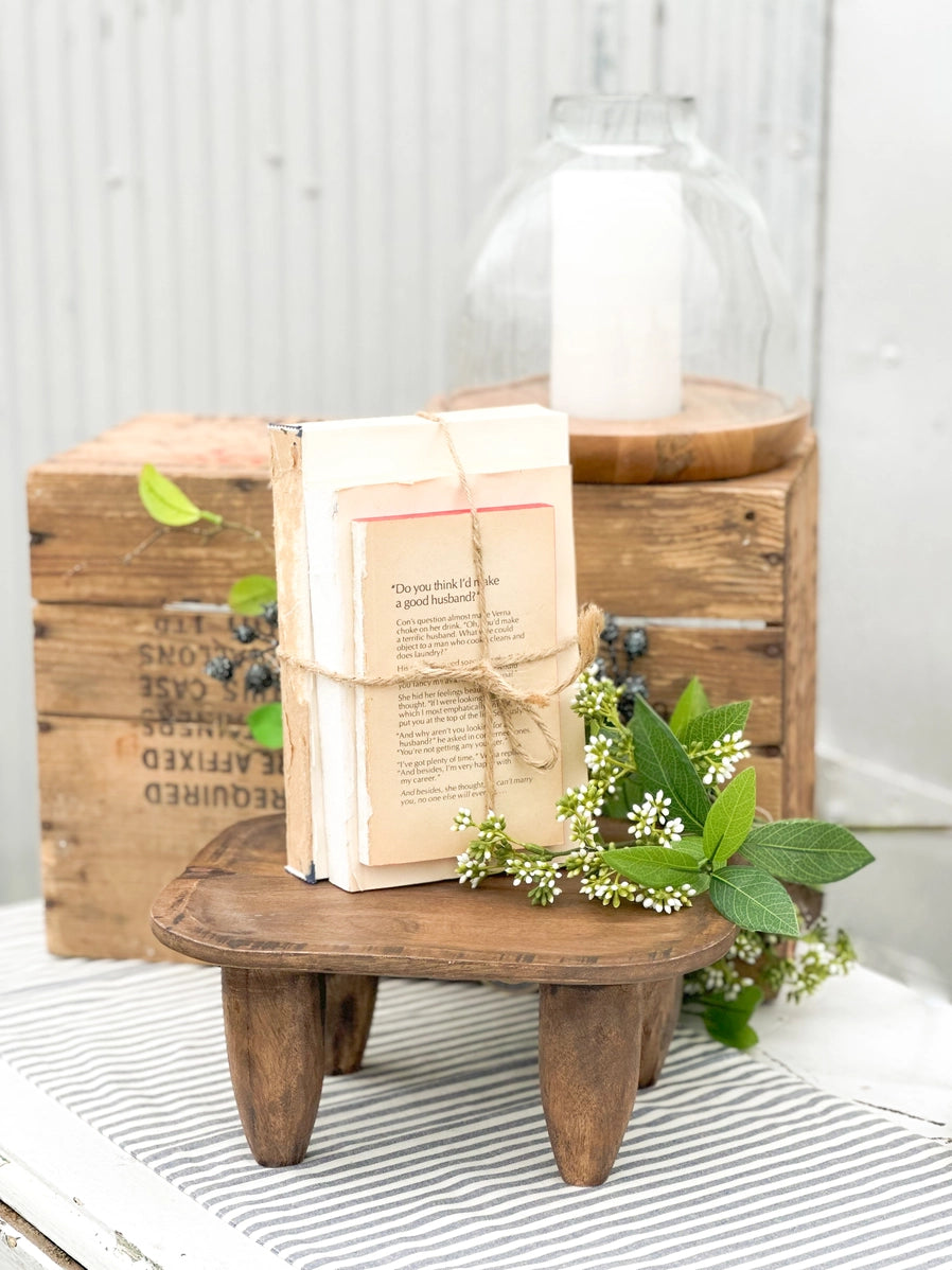 Stack of vintage books wrapped in twine on rustic wooden stool with white flowering greenery and glass cloche backdrop