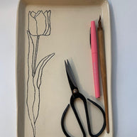 Cream ceramic tray with black line drawing of tulip, containing black scissors, pink pen, and wooden pencil