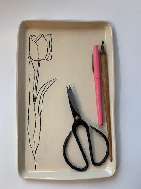 Cream ceramic tray with black line drawing of tulip, containing black scissors, pink pen, and wooden pencil